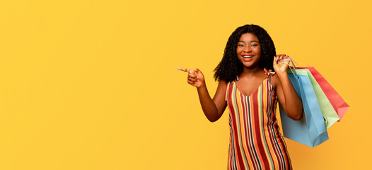 Big sales concept. Portrait of young happy black woman with colorful shopping bags pointing at blank space over orange background, panorama. Excited African American lady enjoying seasonal discounts