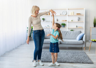 Happy mature lady and casual little daughter dancing waltz at home, standing in living room, full length photo. Loving grandmother teaching granddaughter how to dance having fun together at home