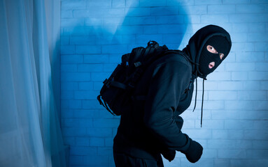 Burglary Concept. Sneaky and scared intruder wearing black balaclava hat lurking in the dark,...