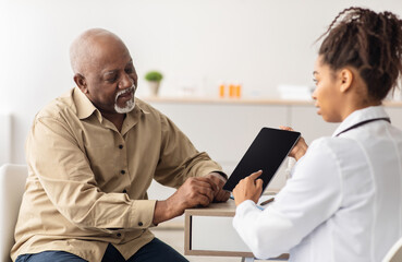 Obraz premium Medicine And Health Care Concept. Black female doctor in white coat holding digital tablet with empty screen for mockup, showing male patient his test results and discussing treatment, sitting at desk