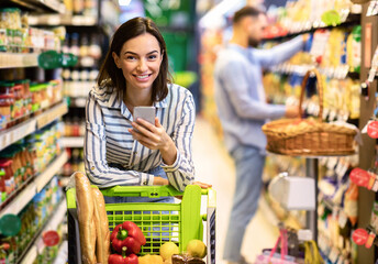 Portrait of smiling young casual woman leaning on shopping trolley cart indoors, using her mobile phone, posing and looking at camera. Satisfied female customer buying products in local grocery store
