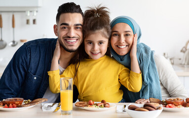 Portrait Of Happy Modern Islamic Family With Little Daughter Having Breakfast Together, Cute Female Child Embracing Parents And Smiling At Camera While Enjoying Tasty Delicious Food In Kitchen