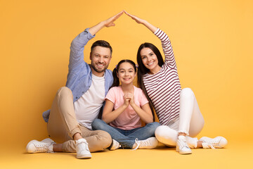 A father and mother are sitting with their daughter in a playful pose as they form a heart shape...