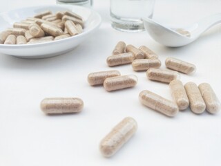 High angle view of capsule pills in a small white plate and in a white spoon, as well as a glass of water and bottle on a white background. 