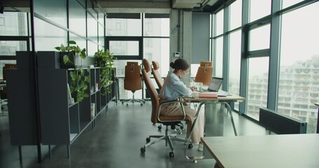 A modern office interior with a focused woman sitting at a desk, working on a laptop and writing notes, surrounded by stylish chairs, shelves with plants, and large glass windows