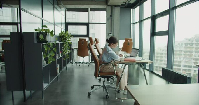 A modern office interior with a focused woman sitting at a desk, working on a laptop and writing notes, surrounded by stylish chairs, shelves with plants, and large glass windows