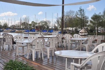 Fototapeta premium Outdoor restaurant tables overlooking a picturesque marina, surrounded by docked boats and calm waters, offering a serene dining experience with a nautical ambiance.