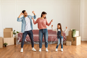 Celebrating Moving Day Concept. Full body length of happy daddy, mum and little girl dancing in new...