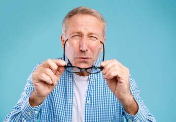 Poor Eyesight. Mature Man Can't See, Squinting Eyes Putting On Glasses Having Problems With Vision, Looking At Camera At Blue Studio. Ophtalmic Issue, Bad Sight In Older Age, Macular Degeneration