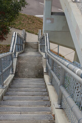 Obraz premium Leading lines looking down tall stairs and landing with metal railings on left beside nature trail in St. Petersburg, FL. Green tree, grass and sidewalk at bottom. Cloudy day. No people room for copy