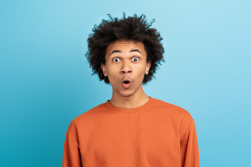 A young African American man displays an expression of surprise with wide eyes and an open mouth, wearing an orange sweater, set against a bright blue backdrop.