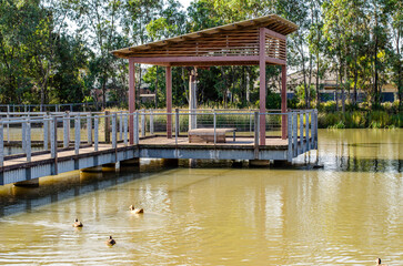 A wooden boardwalk extends into a water pond, leading to a shaded seating area, public shelter or...