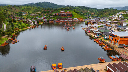 Aerial view of tranquil lakeside village nestled in lush, mist-covered hills under an overcast sky. The calm water reflects the vibrant buildings, raft creating a serene and picturesque scene..