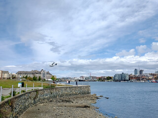 Fototapeta premium A seaplane comes in for a landing on Victoria Harbour with blue sky and white clouds and people walking along a seaside path