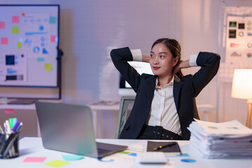 Asian businesswoman relaxing with hands behind head at office desk after finishing work late at...