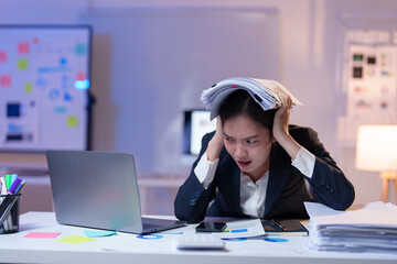 Young stressed businesswoman holding documents on head while working with laptop and financial...