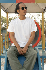 An Asian male model wearing a plain white t-shirt and black sunglasses is posing in a commercial park during the daytime. The male model's white t-shirt is being used as a mockup object for showcasing