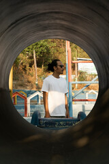 An Asian male model wearing a plain white t-shirt and black sunglasses is posing in a commercial park during the daytime. The male model's white t-shirt is being used as a mockup object for showcasing