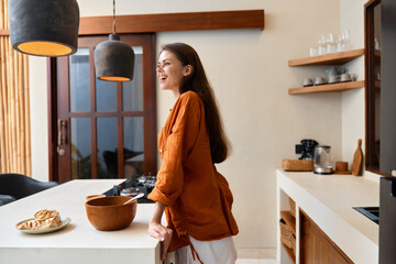 Young woman in a cozy kitchen laughing while preparing food, wearing an orange shirt, expressing...