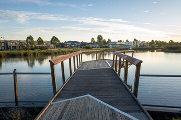 Obraz premium A wooden boardwalk over the still waters of a lake in Tarneit, surrounded by residential houses in the neighborhood. Public recreational amenity in Australia's suburbs. Copy space for design