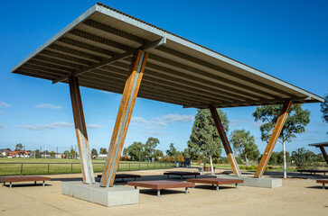 A sleek, modern shelter with a metal roof and wooden supports provides shade in a public space. It...