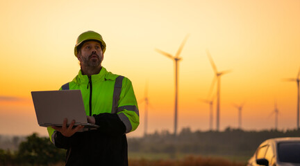 Engineer with laptop survey work in wind turbine farms rotation to generate electricity energy. Maintenance engineer working in wind turbine farm at sunset.