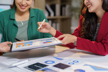 Fototapeta premium Two smiling businesswomen are collaborating on a project, analyzing financial charts and graphs together in a modern office environment, discussing data and strategizing for success
