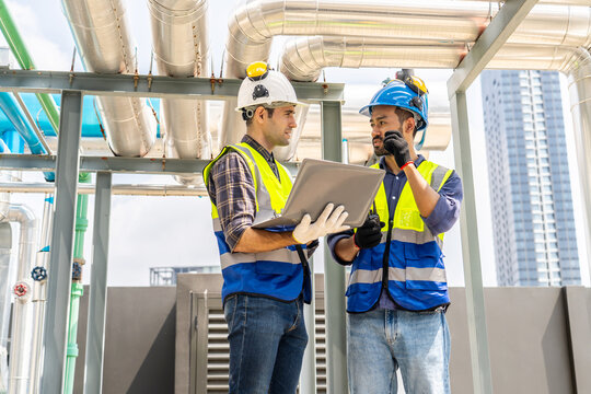 Collaboration, Two young construction workers discuss plans while reviewing blueprints on a rooftop. They wear safety gear and oversee intricate piping systems amidst a bustling urban backdrop,