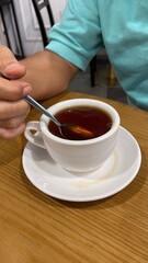 A man’s hand stirring a cup of hot Teh O with a teaspoon on a wooden table, capturing a moment of comfort and relaxation. Ideal for themes of tea culture and Asian cuisine.