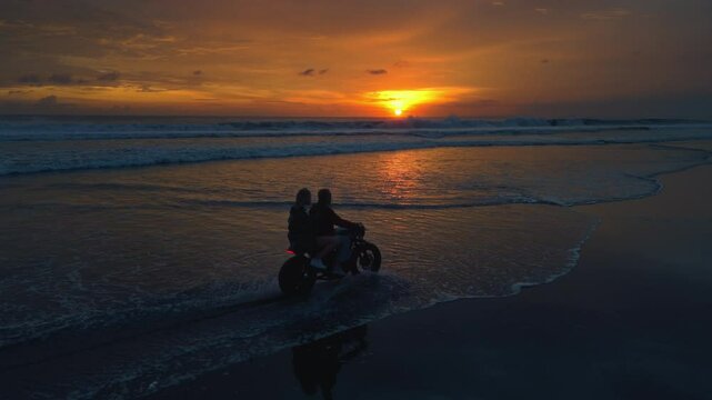 Couple rides retro motorcycle on the beach or shore. Man, woman in love ride a bike in Bali. Trip to ocean or sea at sunset. Travel and road trip. Driving on moped during summer vacation. Aerial drone