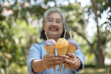  A cheerful elderly woman holding three colorful ice cream cones (purple and orange) extends them...