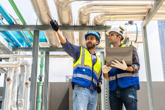 Collaboration, Two young construction workers discuss plans and use laptop computer while reviewing blueprints on a rooftop. They wear safety gear and oversee intricate piping systems in factory
