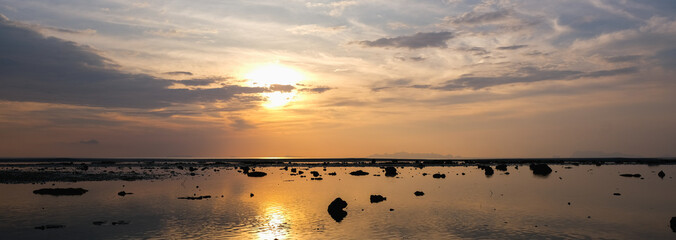 A serene sunset over a calm, reflective sea with scattered rocks and ripples on the water.