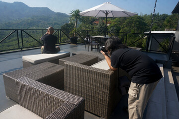 Young Asian Boy Taking Picture Of A Man Sitting On Sofa Using Camera At Terrace Cafe With Mountain View