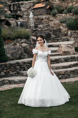 bride in a white wedding dress with a long veil. She stands on a wooden terrace against a background of green trees. The bride holds a small bouquet of white flowers in her hands and smiles.