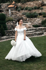 bride in a white wedding dress with a long veil. She stands on a wooden terrace against a background of green trees. The bride holds a small bouquet of white flowers in her hands and smiles.