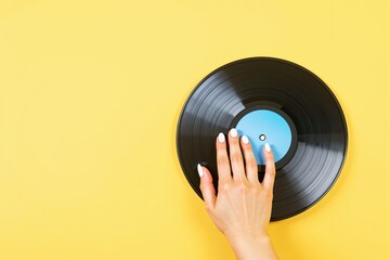 Hand with white nails touching a black vinyl record on yellow background.