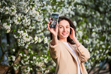 Happy woman taking selfie with smartphone among spring blossoms with soft focus on the background.