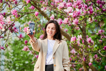 Cheerful woman taking selfie with smartphone among spring blossoms with soft focus on the...