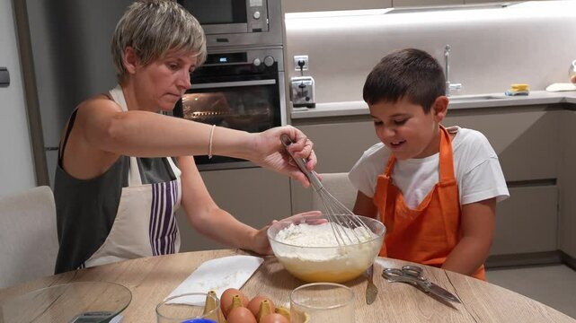 Mom teaching her son how to bake a sponge cake, whisking ingredients in a glass bowl in their modern kitchen