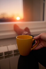 Woman drinking hot tea with her feet in socks on a home heating radiator on a cold winter day outdoors.