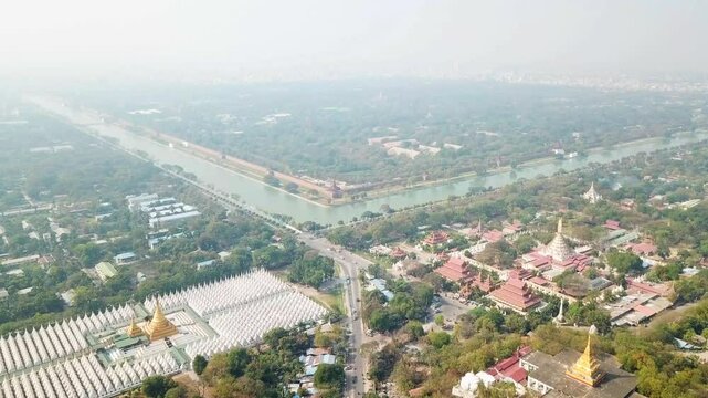 Aerial view captures the cityscape of Mandalay, Myanmar, showcasing a network of waterways, numerous buddhist temples and pagodas, and the dense urban sprawl extending towards the horizon