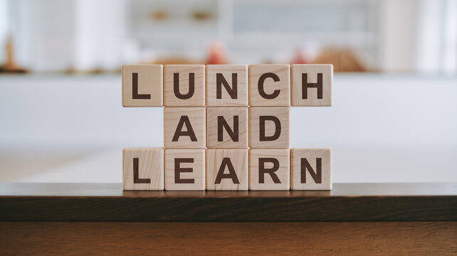 Three wooden blocks stacked vertically on a dark surface read "LUNCH AND LEARN.". A blurred bright background suggests a professional learning setting