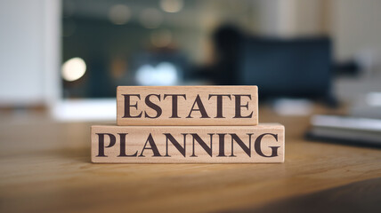 Two wooden blocks stacked on a wooden table display "ESTATE" and "PLANNING" in dark serif font. A blurred indoor background creates a clean, professional focus on estate planning.





