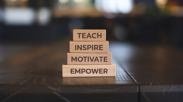 Four stacked wooden blocks display "TEACH," "INSPIRE," "MOTIVATE," and "EMPOWER" on a dark wooden surface. Sharp focus highlights the motivational words against a blurred indoor background.






