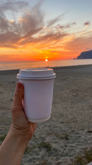 Hand holding a white coffee cup against a stunning beach sunset, capturing the serene moment of enjoying a drink by the ocean during the golden hour.