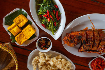 various sundanese traditional food served on wooden table, including fried tilapia fish, water spinach saute, crispy fried dory fish, and sambal dadak