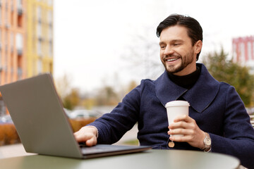 Happy stylish young bearded man holding coffee working with laptop, sitting outdoor in cafe