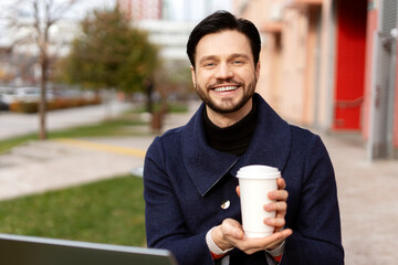 Happy young man in black jacket working with laptop and holding cup with coffee in the city