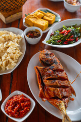 various sundanese traditional food served on wooden table, including fried tilapia fish, water spinach saute, crispy fried dory fish, and sambal dadak